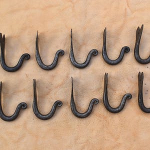 Pode incluir: Dez ganchos de ferro preto forjados à mão dispostos em duas fileiras. Cada gancho apresenta um design curvo com um rolo decorativo e uma extremidade dividida. Os ganchos são exibidos em uma superfície texturizada marrom claro.