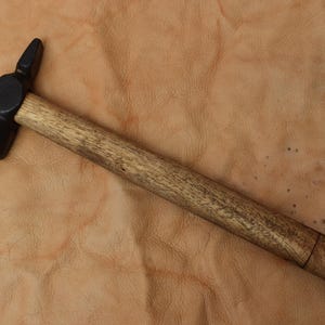 Puede incluir: Un martillo de metal negro con mango de madera. La cabeza del martillo tiene una superficie de golpeo redondeada y una pena puntiaguda. El mango es de color marrón claro y parece ser de madera. El martillo está sobre una superficie de cuero marrón claro.