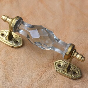 Può includere: Maniglia per armadio in vetro trasparente sfaccettato con finiture in ottone. La maniglia ha una forma a diamante ed è fissata a due placche in ottone ornate con fori per viti. Un accento decorativo per mobili.