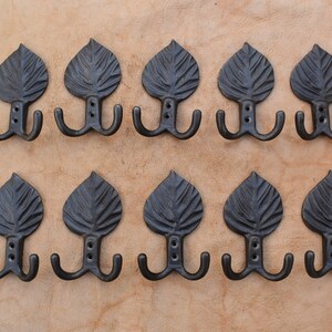 May include: Ten black metal leaf-shaped hooks with two hooks on each. The hooks are mounted on a light brown surface.