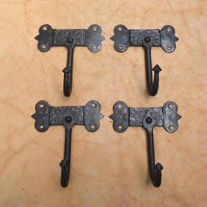 Pode incluir: Um conjunto de quatro ganchos de parede em metal preto, cada um com uma placa traseira retangular martelada com extremidades arredondadas decorativas e quatro orifícios para parafusos. Um gancho vertical central com ponta estende-se de cada placa.