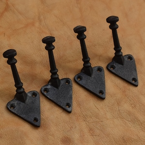 Puede incluir: Cuatro perchas negras de hierro fundido. Cada gancho tiene una parte superior decorativa, un tallo delgado y una base en forma de corazón con agujeros pretaladrados para el montaje. Los ganchos están dispuestos en fila sobre un fondo de cuero marrón.