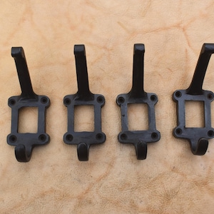 Pode incluir: Quatro ganchos de parede em metal preto dispostos em fila. Cada gancho apresenta uma estrutura retangular com quatro orifícios para parafusos, um gancho longo curvado para cima e um gancho menor curvado para baixo na base.