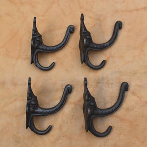 May include: Four black cast iron wall hooks with an elephant head design. The hooks are arranged in a square pattern against a tan background. Each hook has a curved arm for hanging items.