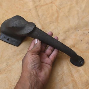 May include: A dark brown cast iron door handle with an elephant head design. The handle is held against a tan leather surface. The handle has a rectangular base with two screw holes and a curved, elongated grip.