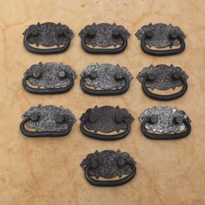 May include: A set of ten dark gray metal drawer pulls arranged in a grid. Each pull features a textured, oval-shaped backplate with scalloped edges and a curved, swinging handle attached by two circular mounting points on the front surface.