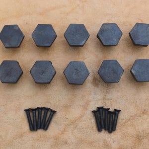 Könnte beinhalten: Ein Set aus zehn dunkelgrauen, sechseckigen Knöpfen mit flacher Oberseite, angeordnet in zwei Reihen zu je fünf Stück. Unter den Knöpfen befinden sich zehn kleine schwarze Metallschrauben mit Flachkopf, sortiert in zwei Fünfergruppen.