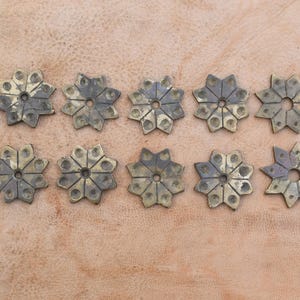 May include: Ten decorative metal star-shaped embellishments with a textured, aged finish. The stars are arranged in two rows of five on a light brown surface.