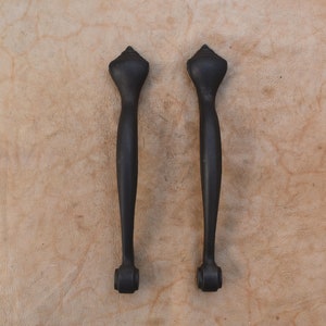 May include: Two black metal door pulls with a pointed top and a rounded bottom. The pulls are set against a light brown background.