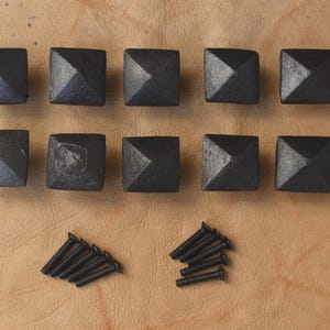 May include: Ten square, black metal cabinet knobs with a pyramid design, arranged in two rows. Two sets of black screws are below the knobs. The knobs and screws are on a tan leather surface.