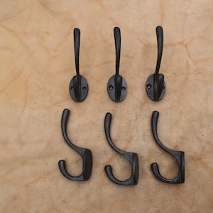 Pode incluir: Um conjunto de seis ganchos de parede de metal preto dispostos em duas filas de três. Cada gancho apresenta um design curvo com uma base de montagem circular contendo dois orifícios para parafusos. Os ganchos têm um acabamento escuro e liso.