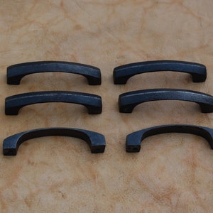May include: Six black metal cabinet pulls with a rounded rectangular shape. The pulls are arranged in two rows of three.