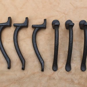 Könnte beinhalten: Ein Set aus sechs schwarzen Metall-Schrankgriffen, angeordnet in zwei Dreiergruppen. Die ersten drei Griffe haben ein geschwungenes, glattes Design, während die anderen drei Griffe eine texturierte, gehämmerte Oberfläche und eine gerade Form aufweisen.