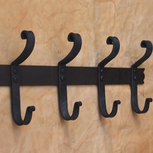 May include: A black, wrought iron wall-mounted coat rack with four curved hooks. The rack has a horizontal bar with decorative edges and is mounted on a light brown surface. The hooks are evenly spaced.