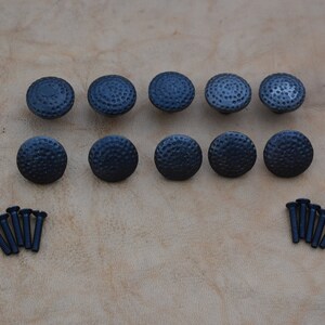 May include: Ten black metal cabinet knobs with a hammered finish and a round shape. The knobs are arranged in two rows of five. The image also shows ten black screws.
