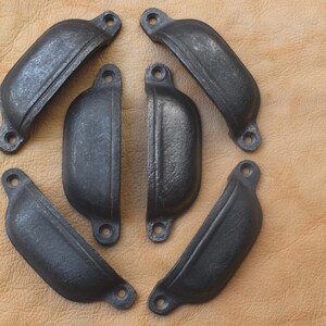 May include: Six dark gray cast iron cup pulls arranged on a tan leather surface. Each pull has a curved shape with two mounting holes. The pulls have a rustic, aged appearance, suggesting a vintage or industrial style.