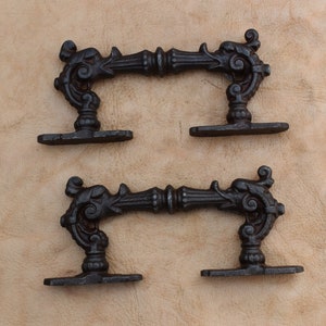 Op de afbeelding: Twee sierlijke, zwarte gietijzeren handgrepen met een krulontwerp. Elke handgreep heeft een centrale balk met decoratieve krullen en twee montageplaten. De handgrepen worden weergegeven tegen een getextureerde, bruinachtige achtergrond.