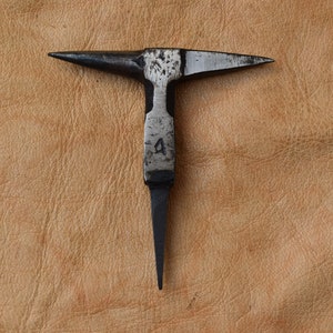Puede incluir: Un yunque de metal negro con una cabeza en forma de T y un extremo puntiagudo. El yunque tiene el número "4" estampado en el eje. La herramienta está sobre un fondo de cuero marrón claro.