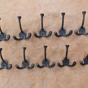 Könnte beinhalten: Zwölf schwarze Metallhaken mit jeweils drei Zacken, angeordnet in zwei Reihen zu sechs auf einer hellbraunen Oberfläche.