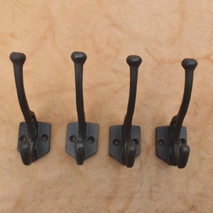 Pode incluir: Um conjunto de quatro ganchos de parede de metal castanho escuro, cada um com um design de ponta dupla com um gancho superior longo e um gancho inferior mais curto. Cada gancho tem uma placa de montagem plana com dois orifícios para parafusos.