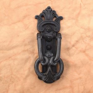 May include: Ornate black door knocker with a decorative floral design. The knocker features a ring pull and a detailed, symmetrical design. The hardware is made of metal and is mounted on a light brown surface.