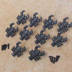 Pode incluir: Dez puxadores de gaveta de lagosta de ferro fundido preto com acabamento rústico. Os puxadores estão dispostos em círculo sobre uma superfície marrom claro. Os puxadores vêm com dez parafusos pretos.