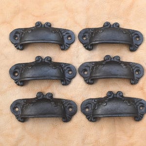 May include: Six antique-style, black metal drawer pulls arranged on a light brown surface. Each pull has a curved handle with decorative scrollwork and two mounting holes. The pulls are likely for furniture restoration.