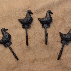 May include: Four black cast iron duck hooks. Each hook is shaped like a duck with a detailed design. The hooks are mounted on a brown surface.