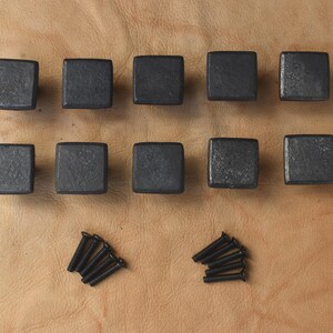 May include: Ten square, dark gray cabinet knobs with a textured surface are arranged on a light brown surface. Two sets of screws are also present. These knobs are likely for furniture or cabinetry.