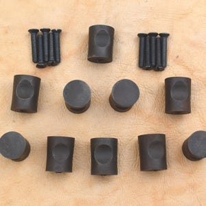 Peut inclure: Un ensemble de quincaillerie d'armoire gris foncé, comprenant des boutons cylindriques et des poignées en forme de barillet, ainsi que des vis assorties. L'ensemble est disposé sur une surface brun clair.
