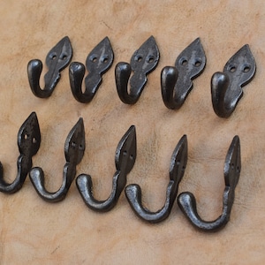 May include: Eight dark grey metal hooks are displayed. Each hook features a decorative leaf-shaped top with two small holes and a curved hook. The hooks are arranged in two rows on a light brown surface.