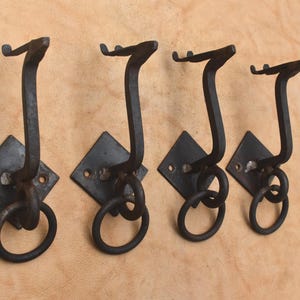 May include: A set of four dark gray metal wall hooks, each featuring a diamond-shaped mounting plate with two screw holes, a curved upward-facing hook arm, and a circular ring attached to the base of the hook for hanging various items.