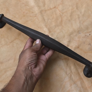 May include: A dark, matte black door pull handle. The handle is held by a hand and is set against a tan background. The handle is approximately 12 inches long and has a geometric design.