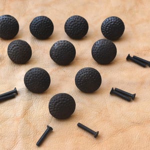 Könnte beinhalten: Zehn schwarze, runde Schrankknöpfe mit strukturierter Oberfläche, die Golfbällen ähneln, sind auf einer hellbraunen Oberfläche angeordnet. Jeder Knopf wird von einer schwarzen Schraube begleitet. Diese dekorativen Knöpfe sind für Möbel geeignet.