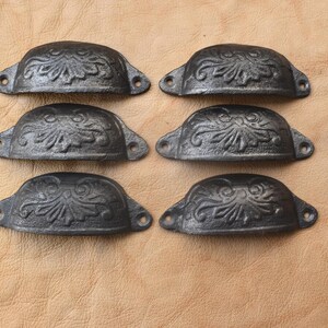 May include: Six antique-style, dark gray metal cup pulls with a floral design. Each pull has two holes for mounting. The pulls are arranged on a light brown leather surface.