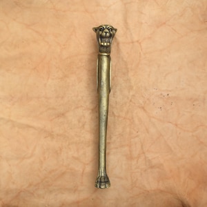 May include: A metallic gold-toned object featuring a sculpted feline head at the top with an open mouth showing teeth. The elongated, slender body tapers downward to a base shaped like a paw with visible claws, finished in a textured, matte metal.