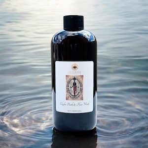 May include: A dark brown bottle with a black cap, labeled "Elegba Bath & Floor Wash." The label features an illustration of a figure and the text "16 FL OZ/473 mL." The bottle is set against a water background.