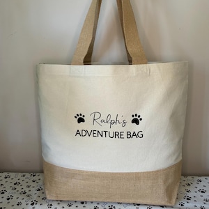 May include: A beige canvas tote bag with brown jute trim. The bag has black paw prints and the text "Ralph's Adventure Bag" printed on the front.