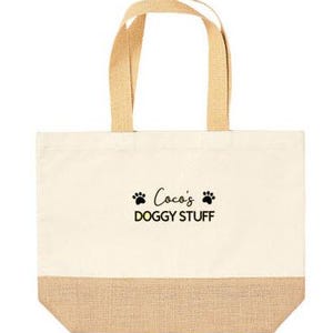 May include: A beige canvas tote bag with brown jute trim. The bag has two brown fabric handles and features black text that reads "Coco's Doggy Stuff" with two black paw prints on either side of the text.