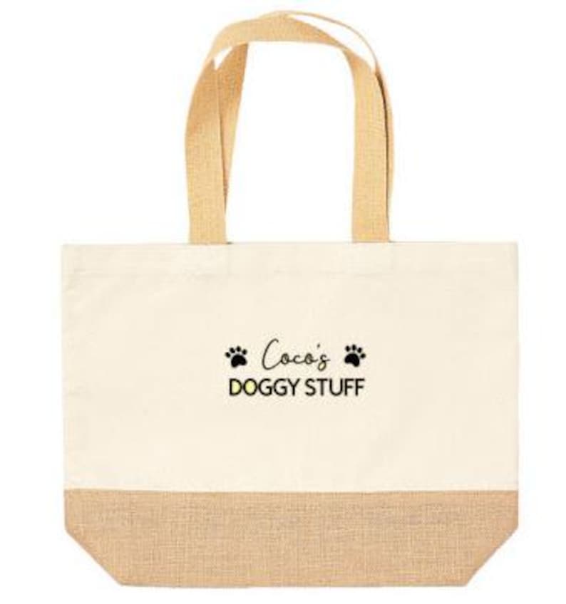 May include: A beige canvas tote bag with brown jute trim. The bag has two brown fabric handles and features black text that reads "Coco's Doggy Stuff" with two black paw prints on either side of the text.