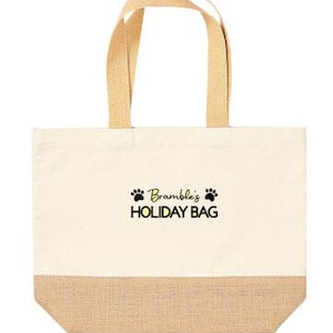May include: A beige canvas tote bag with a brown jute bottom. The bag has black paw prints and the text "Bramble's HOLIDAY BAG" in yellow.