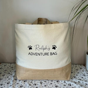 May include: A beige canvas tote bag with a brown jute bottom. The bag has black paw prints and the text "Ralph's Adventure Bag" printed on the front.