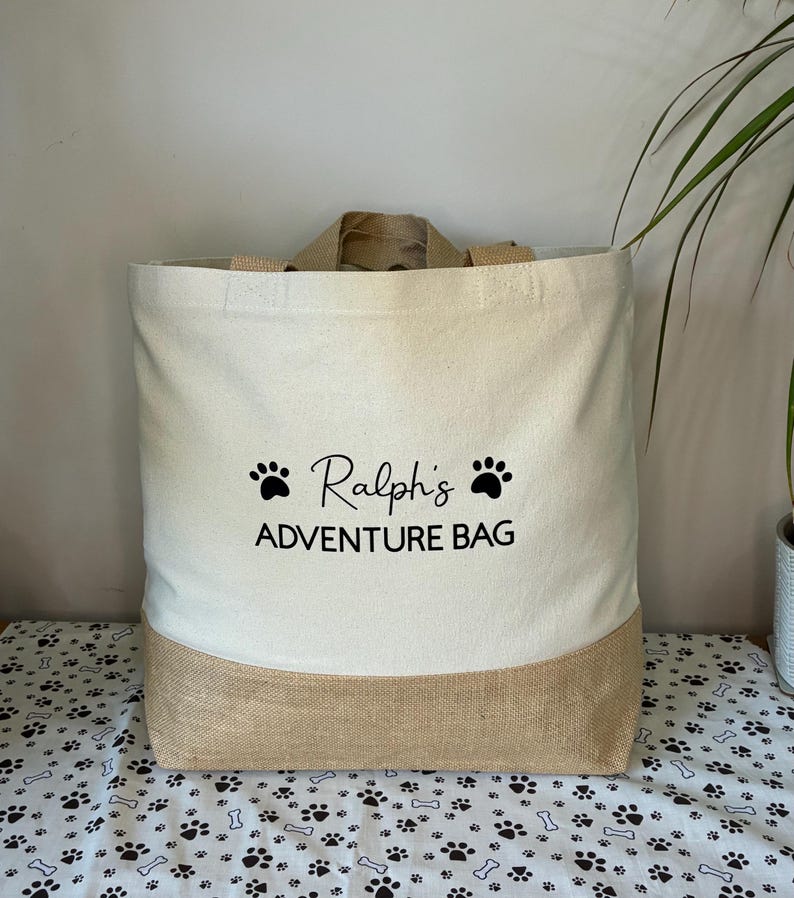 May include: A beige canvas tote bag with a brown jute bottom. The bag has black paw prints and the text "Ralph's Adventure Bag" printed on the front.