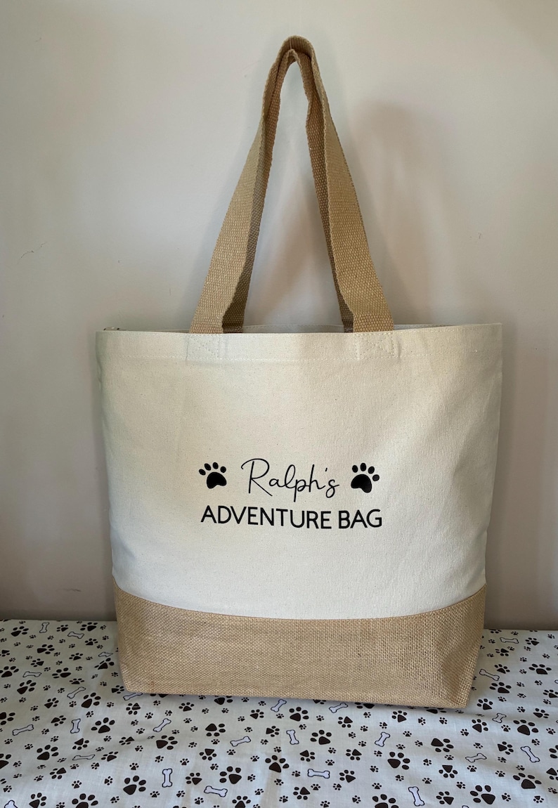 May include: A beige canvas tote bag with brown jute trim. The bag has black paw prints and the text "Ralph's Adventure Bag" printed on the front.