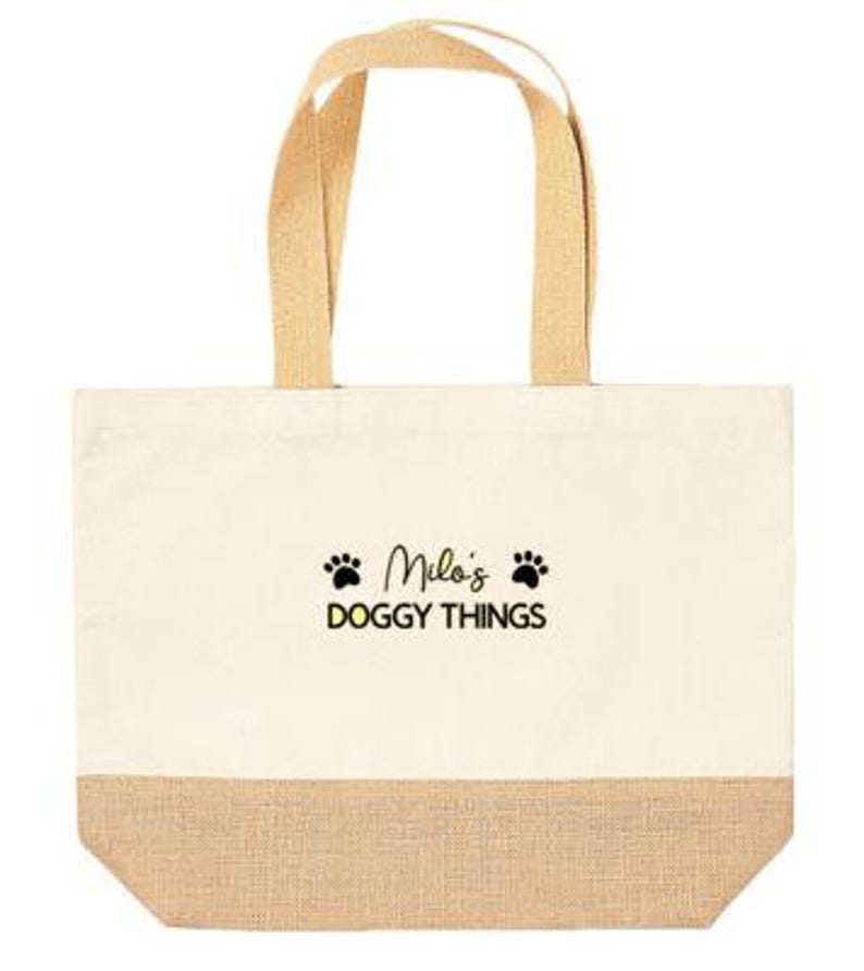 May include: A beige canvas tote bag with brown jute trim. The bag has two brown fabric handles and features black paw prints and the text "Milo's Doggy Things" in black.