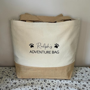 May include: A beige canvas tote bag with brown jute trim. The bag has black paw prints and the text "Ralph's Adventure Bag" printed on the front.