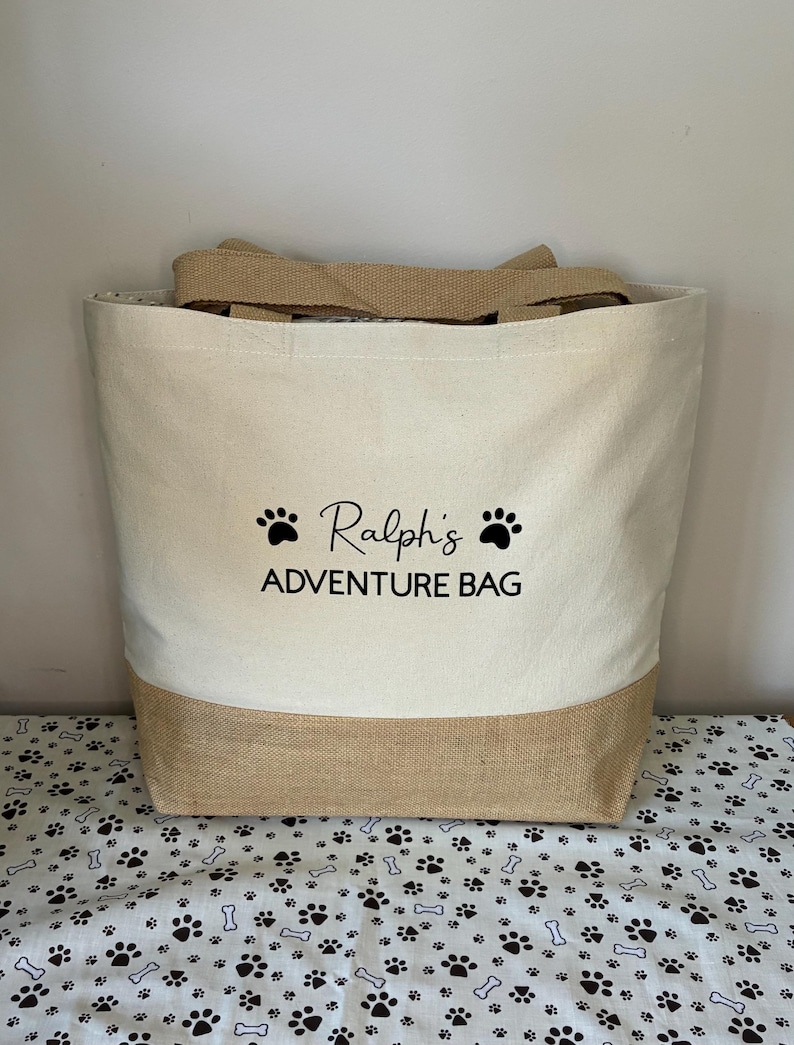 May include: A beige canvas tote bag with brown jute trim. The bag has black paw prints and the text "Ralph's Adventure Bag" printed on the front.