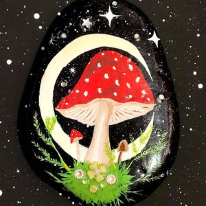 May include: Hand-painted rock art featuring a red and white mushroom with white spots, surrounded by green foliage and a crescent moon on a black background with white stars.