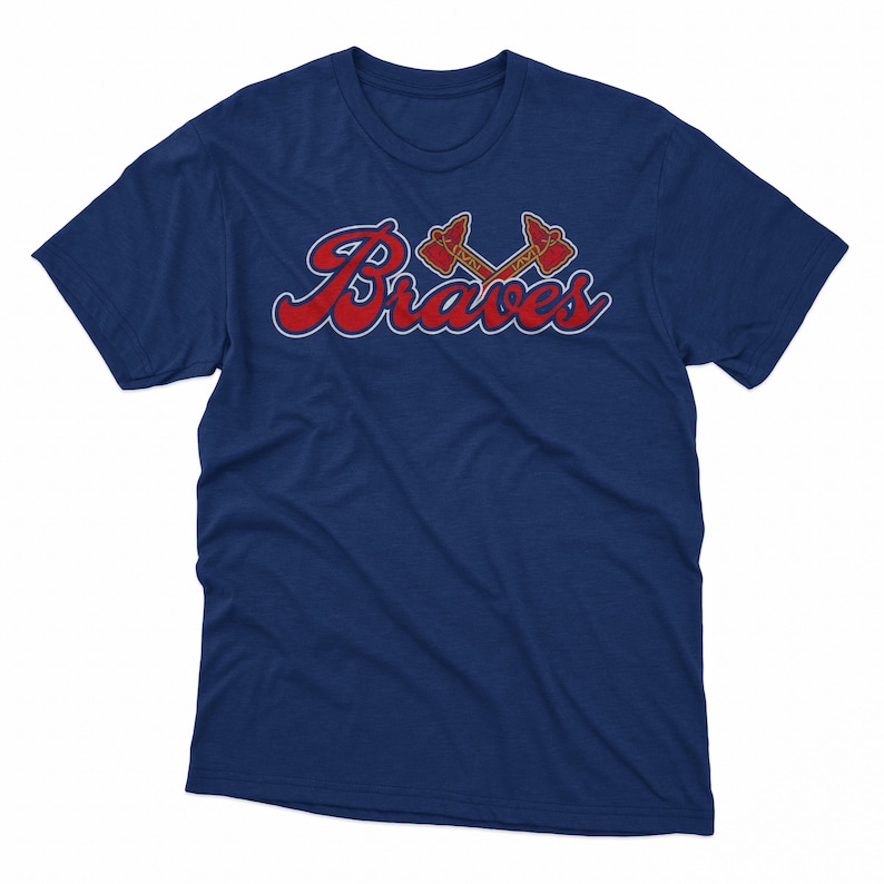 Puede incluir: Una camiseta azul marino con la palabra "Braves" en escritura roja y un contorno blanco. Encima de la palabra hay dos tomahawks cruzados en rojo y dorado. La camiseta tiene cuello redondo y mangas cortas.