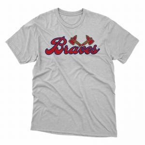 Puede incluir: Camiseta gris claro con la palabra "Braves" en rojo con un contorno azul. Encima de la palabra hay dos tomahawks cruzados en rojo y dorado.